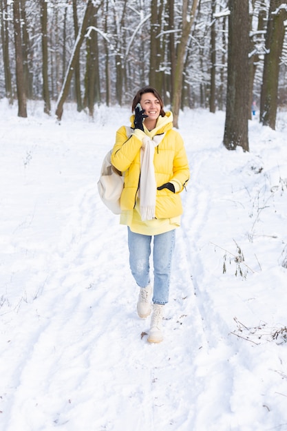 Glückliche Frau in einer guten Stimmung geht durch den verschneiten Winterwald und plaudert fröhlich am Telefon, genießt Zeit draußen im Park