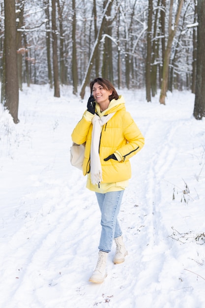 Glückliche Frau in einer guten Stimmung geht durch den verschneiten Winterwald und plaudert fröhlich am Telefon, genießt Zeit draußen im Park