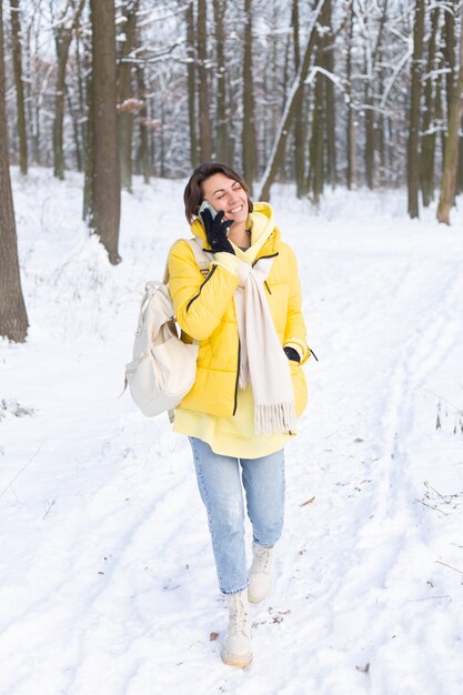 Glückliche Frau in einer guten Stimmung geht durch den verschneiten Winterwald und plaudert fröhlich am Telefon, genießt Zeit draußen im Park