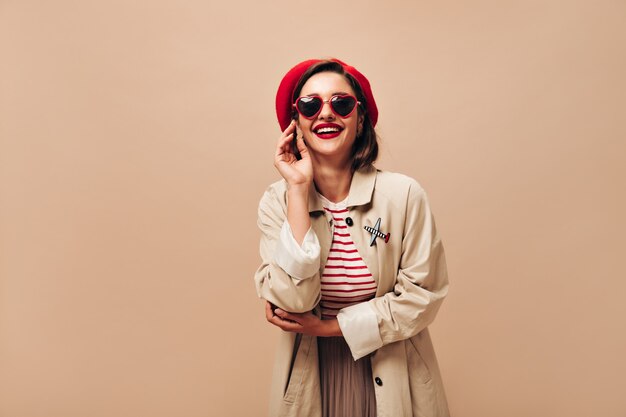 Glückliche Frau in Sonnenbrille, Hut und Graben lächelt auf lokalisiertem Hintergrund. Freudige Dame im gestreiften Pullover und im beigen Mantel, der aufwirft.