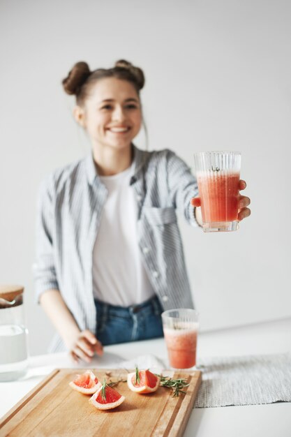 Glückliche Frau mit Brötchen lächelnd, das Glas mit Grapefruit-Detox-Smoothie zu jemandem streckt. Weißer Wandhintergrund. Gesunde Ernährung.