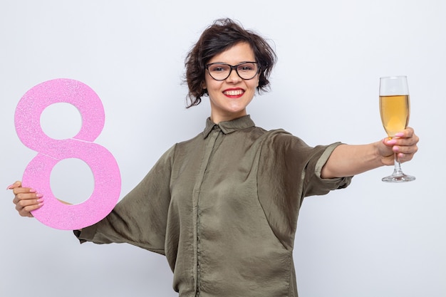 Glückliche frau mit den kurzen haaren, die nummer acht halten, hergestellt aus pappe und einem glas champagner lächelnd, das fröhlich internationalen frauentag 8. märz feiert