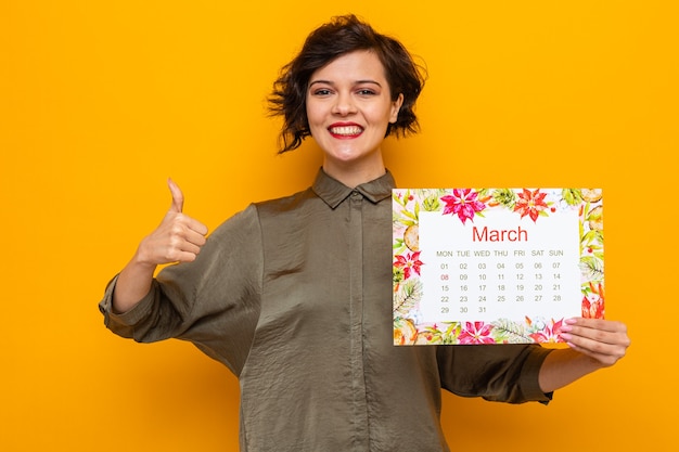 Glückliche Frau mit kurzen Haaren, die den Papierkalender des Monats März hält und die Kamera anschaut, die fröhlich lächelt und Daumen nach oben zeigt, um den internationalen Frauentag 8. März zu feiern, der über orangefarbenem Hintergrund steht