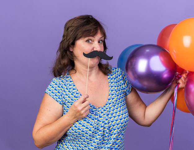 Glückliche Frau mittleren Alters, die bunte Luftballons und lustigen Schnurrbart am Stock hält und Spaß hat having