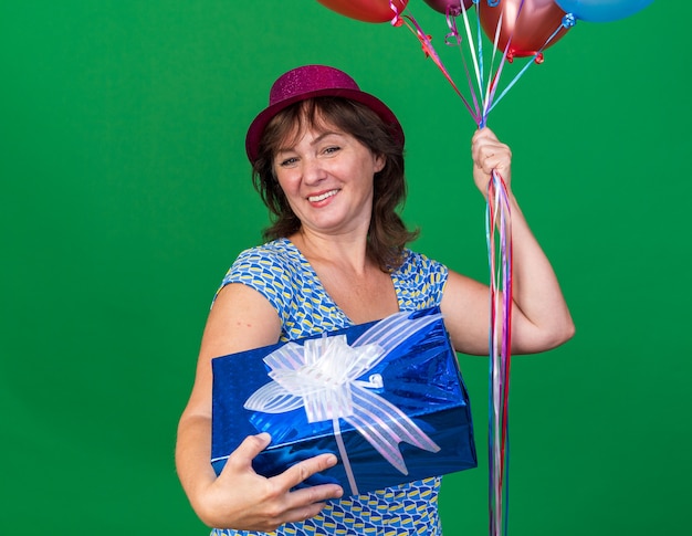 Glückliche Frau mittleren Alters mit Partyhut, die bunte Luftballons hält und fröhlich lächelnd die Geburtstagsfeier feiert, die über grüner Wand steht