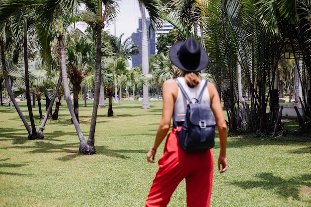 Glückliche Frau reisen durch Bangkok mit Rucksack und genießen schönen sonnigen Tag im tropischen Park auf grünem Grasfeld