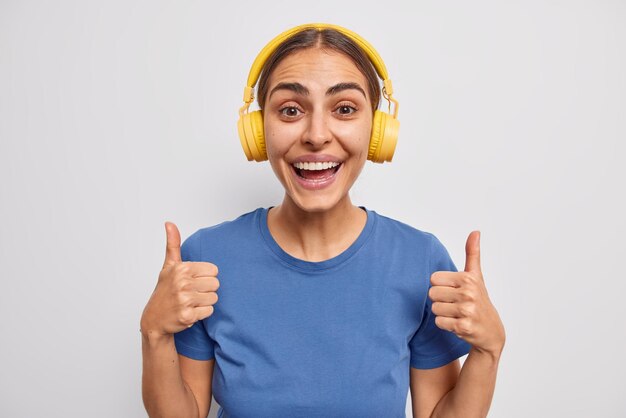 Glückliche Frau stimmt zu, dass etwas die Daumen hochhält, empfiehlt, diese Audiospur anzuhören, trägt gelbe Stereokopfhörer auf den Ohren, die in einem blauen T-Shirt isoliert auf weißem Hintergrund gekleidet sind. Ich mag das.