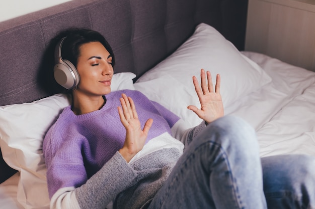 Glückliche Frau zu Hause auf bequemem Bett, das warmen Kleiderpullover trägt, Musik hören