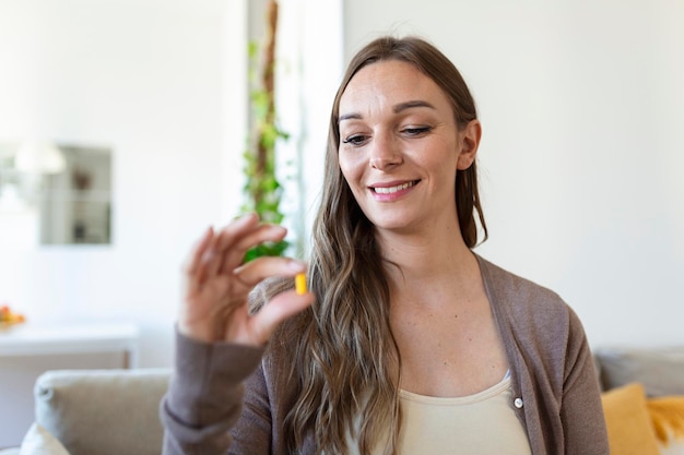 Glückliche frau zu hause, die zu hause am tisch und in der küche sitzt und eine tasse wasser und eine tabletten-coronavirus-behandlung hält, nahaufnahme, unscharfer hintergrund