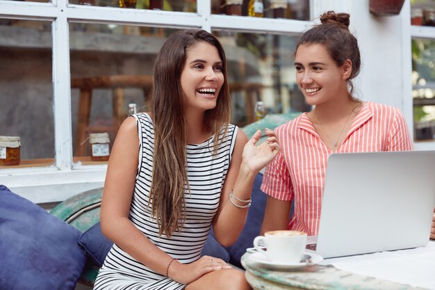 Glückliche Frauen im Café, haben Spaß zusammen, surfen im Internet auf einem Laptop, genießen heißen aromatischen Cappuccino, haben positive Ausdrücke. Menschen, Freizeit und Freundschaftskonzept.