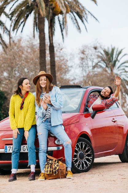 Glückliche Frauen mit Smartphone nahe dem Mann, der sich heraus vom Auto lehnt