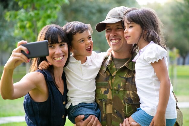 Glückliche freudige Militärfamilie, die Väter feiert, die zurückkehren, Freizeit im Park genießen, Selfie auf Smartphone nehmen. Mittlerer Schuss. Familientreffen oder Rückkehr nach Hause Konzept