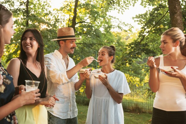 Glückliche Freunde essen und trinken Bier beim Grillabendessen bei Sonnenuntergang