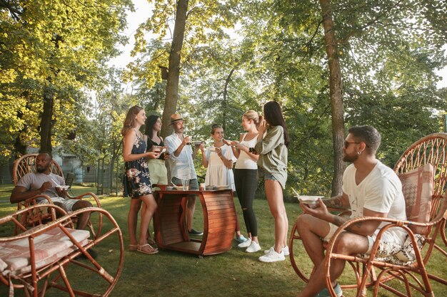 Glückliche Freunde essen und trinken Bier beim Grillabendessen bei Sonnenuntergang