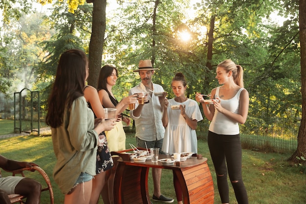 Glückliche Freunde essen und trinken Bier beim Grillabendessen bei Sonnenuntergang