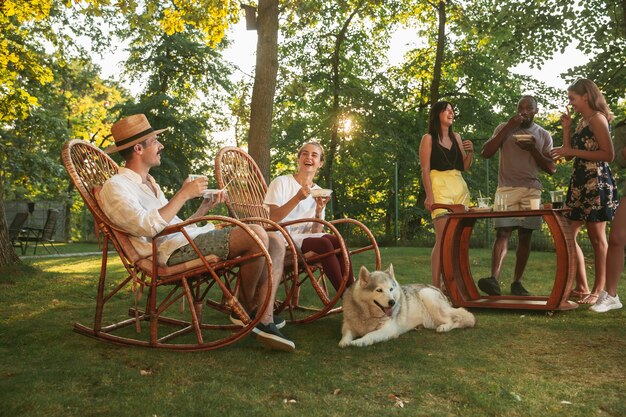 Glückliche Freunde essen und trinken Bier beim Grillabendessen bei Sonnenuntergang