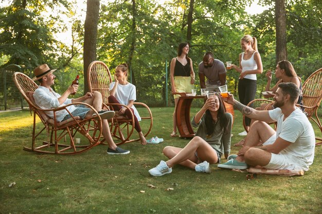 Glückliche Freunde essen und trinken Bier beim Grillabendessen bei Sonnenuntergang