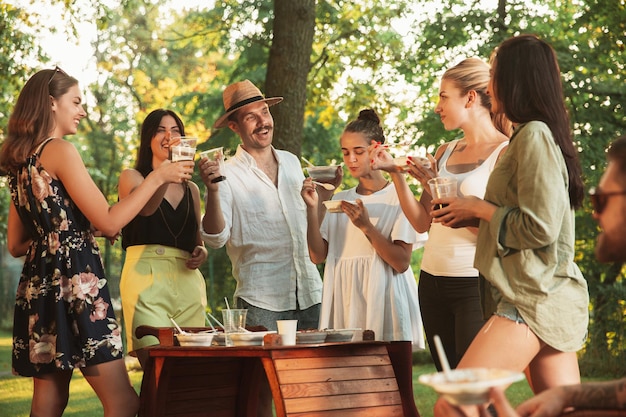 Glückliche Freunde essen und trinken Bier beim Grillabendessen bei Sonnenuntergang