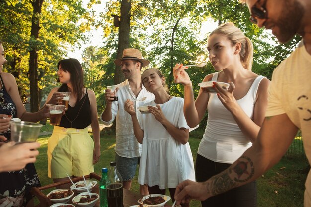 Glückliche Freunde essen und trinken Bier beim Grillabendessen bei Sonnenuntergang