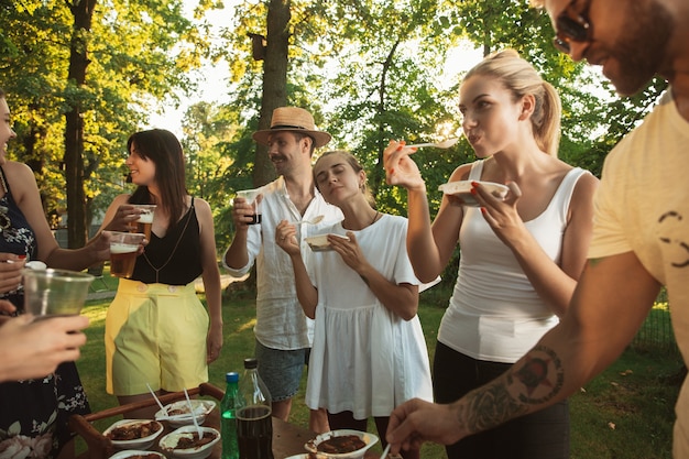 Glückliche Freunde essen und trinken Bier beim Grillabendessen bei Sonnenuntergang