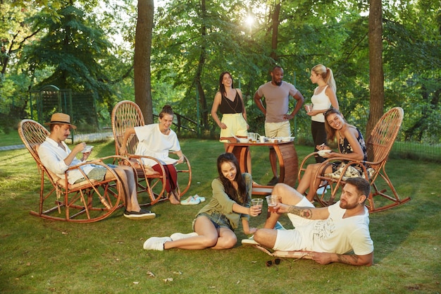 Glückliche Freunde essen und trinken Bier beim Grillabendessen bei Sonnenuntergang