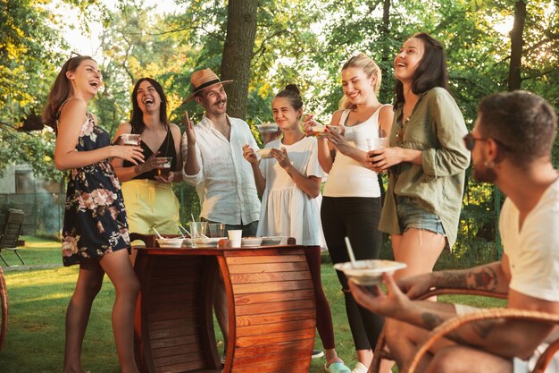 Glückliche Freunde essen und trinken Bier beim Grillabendessen bei Sonnenuntergang