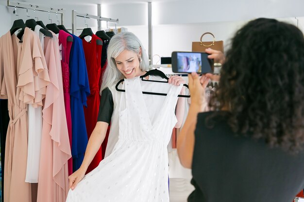 Glückliche Freundinnen genießen das Einkaufen im Bekleidungsgeschäft zusammen, halten Kleid, posieren und fotografieren auf Handy. Konsum- oder Einkaufskonzept