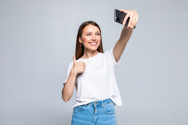 Glückliche fröhliche junge Frau, die Daumen oben zeigt und Selfie auf Smartphone lokalisiert macht