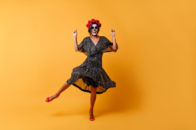 Glückliche gebräunte Frau hat Spaß beim Tanzen im Halloween-Bild. In voller Länge Aufnahme eines Mädchens im schwarzen Outfit und mit Rosen im Haar
