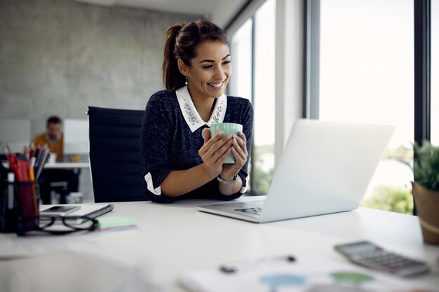 Glückliche Geschäftsfrau, die während ihrer Kaffeepause im Büro einen Videoanruf über Laptop tätigt