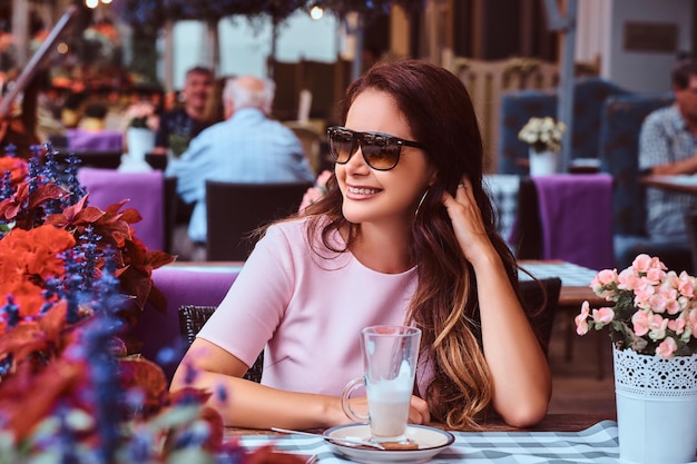 Glückliche Geschäftsfrau mittleren Alters mit langen braunen Haaren, die ein rosafarbenes Kleid trägt und mit einem Glas Cappuccino im Café im Freien sitzt.