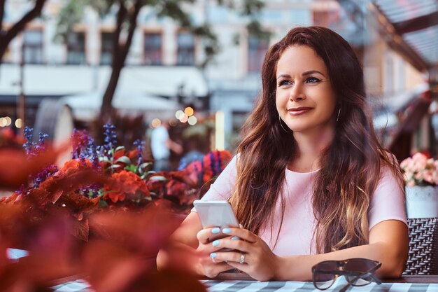 Glückliche Geschäftsfrau mittleren Alters mit langen braunen Haaren hält ein Smartphone in der Hand, während sie in einem Café im Freien sitzt.