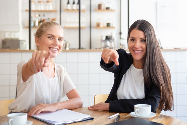 Glückliche Geschäftsfrauen, die Finger auf Kamera zeigen und zeigen, während sie am Tisch mit Kaffeetassen und Dokumenten sitzen