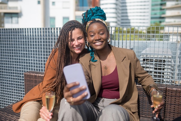 Glückliche Geschäftsfrauen, die Selfie auf der Terrassendachparty machen. Frauen in formeller Kleidung sitzen auf einem Rattansofa, schauen auf das Handy und lächeln. Teambuilding, Party, Social Media Konzept