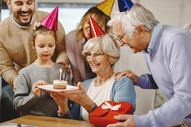 Glückliche Großfamilie, die den Geburtstag der Seniorin feiert und sie mit einem Kuchen und Geschenken überrascht. Der Fokus liegt auf der Seniorin