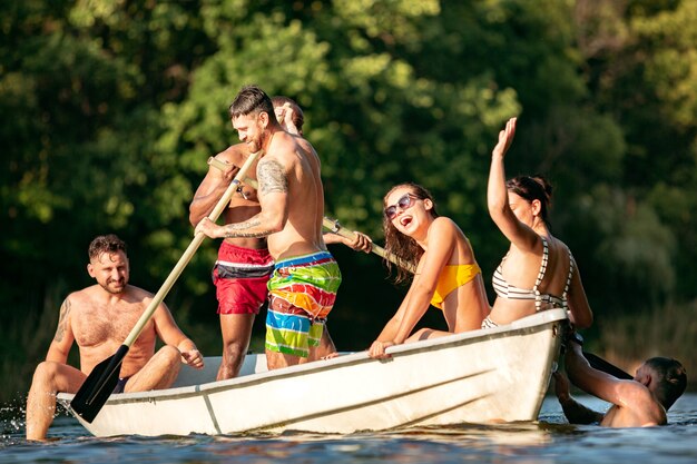 Glückliche Gruppe von Freunden, die Spaß beim Lachen und Schwimmen im Fluss haben. Freudige Männer und Frauen im Badeanzug in einem Boot am Flussufer an sonnigem Tag. Sommerzeit, Freundschaft, Resort, Wochenendkonzept.