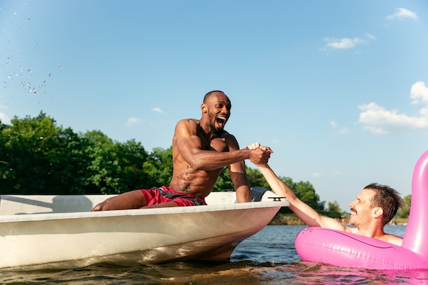 Glückliche Gruppe von Freunden, die Spaß haben, während sie lachen, Wasser spritzen und im Fluss schwimmen. Freudige Männer im Badeanzug in einem Boot am Flussufer an sonnigem Tag. Sommerzeit, Freundschaft, Resort, Wochenendkonzept.