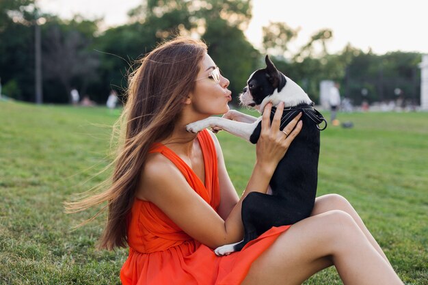 Glückliche hübsche Frau, die auf Gras im Sommerpark sitzt, Boston-Terrier-Hund hält, küsst, orange Kleid trägt, trendigen Stil, spielt mit Haustier