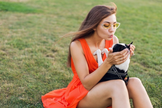 Glückliche hübsche Frau, die auf Gras im Sommerpark sitzt, Boston-Terrier-Hund hält, küsst, orange Kleid trägt, trendigen Stil, spielt mit Haustier