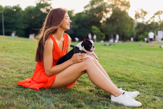 Glückliche hübsche Frau, die auf Gras im Sommerpark sitzt, Boston-Terrier-Hund hält, positive Stimmung lächelt, orange Kleid trägt, trendigen Stil, schlanke Beine, Turnschuhe, spielt mit Haustier
