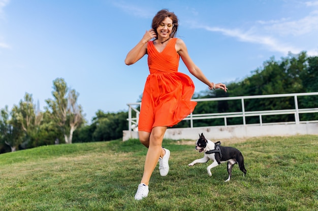 Glückliche hübsche Frau im Park, der mit Boston-Terrier-Hund läuft, positive Stimmung lächelt, trendiger Sommerstil, orange Kleid trägt, mit Haustier spielt, Spaß hat, bunte, aktive Wochenendferien, Turnschuhe