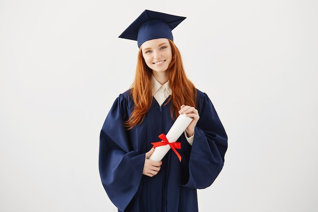 Glückliche Ingwer-Absolventin in der Kappe und im Mantel lächelnd, die Diplom halten.