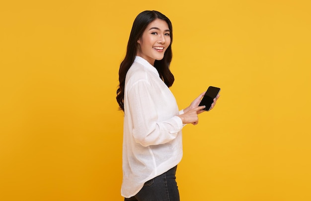 Glückliche junge asiatische Frau, die am Handy des leeren Bildschirms lokalisiert über gelbem Hintergrund zeigt
