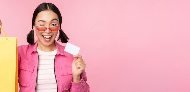 Glückliche junge asiatische frau, die kreditkarte für den einkauf zeigt, der den taschenkauf im verkauf hält, der zum shop geht, der über rosa hintergrund steht