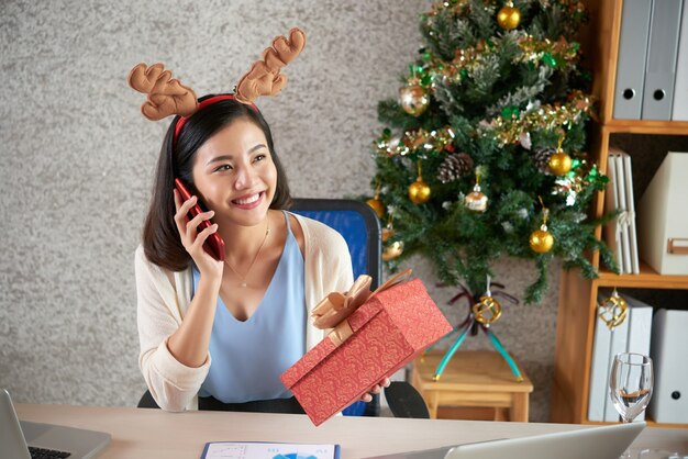 Glückliche junge asiatische Frau im Rentierhaarband, das Weihnachtsgeschenk hält und am Telefon spricht