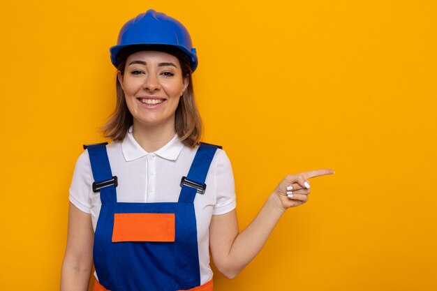 Glückliche junge Baumeisterin in Bauuniform und Schutzhelm, die fröhlich lächelt und mit dem Zeigefinger zur Seite zeigt