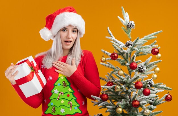 Glückliche junge blonde Frau im Weihnachtspullover und in der Weihnachtsmannmütze, die ein Geschenk hält, das Kamera betrachtet, das dankbar steht, neben einem Weihnachtsbaum über orange Hintergrund zu stehen