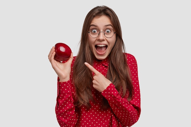 Glückliche junge Dame zeigt auf roten saftigen Apfel, zeigt gesundes Essen, hält sich an Diät, hält Kiefer herunter, hat langes Haar, erstaunten Gesichtsausdruck