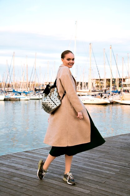 Glückliche junge elegante Frau, die am Luxusyachtclub von Barcelona geht, Mantel Turnschuhe und Rucksack tragend, touristische Zeit der Zwischensaison.