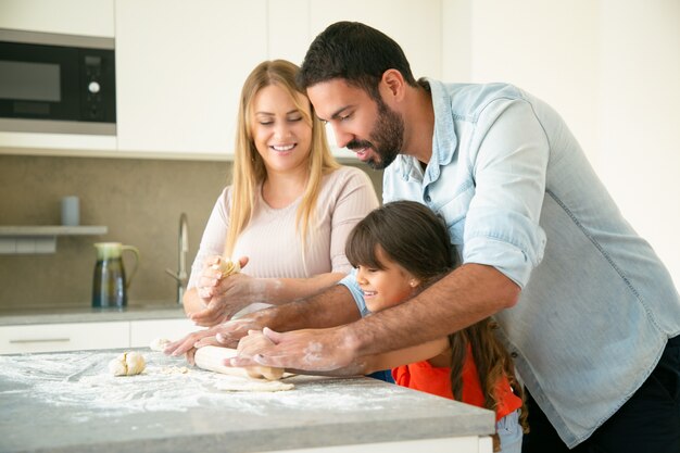 Glückliche junge Eltern, die Tochter lehren, Teig auf Küchenschreibtisch mit Mehl unordentlich zu rollen. Junges Paar und ihr Mädchen backen Brötchen oder Kuchen zusammen. Familienkochkonzept